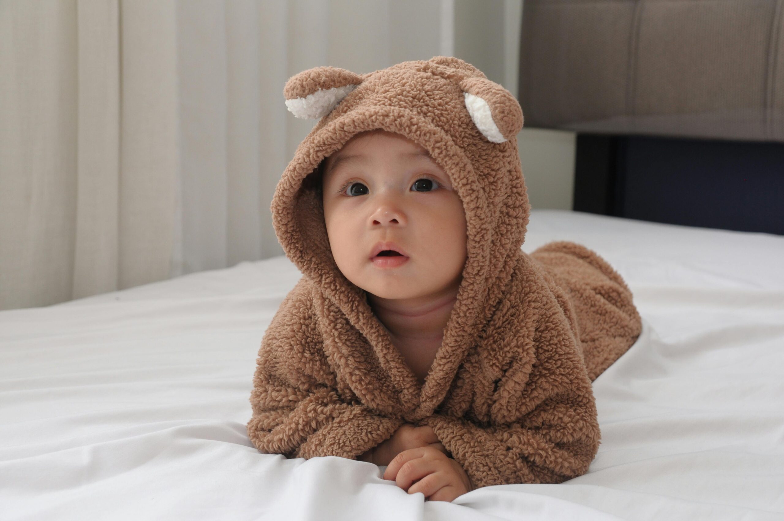 Adorable baby in bear costume on white bed, exuding cuteness and warmth indoors.